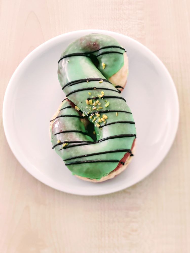 A doughnot in the shape of the Mahara logo on a plate. The donout has green glazing with horizontal chocolate stripes